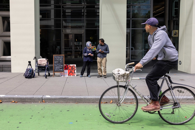 A person wearing a helmet rides a bicycle on a path in front of a building with large windows and some outdoor seating.